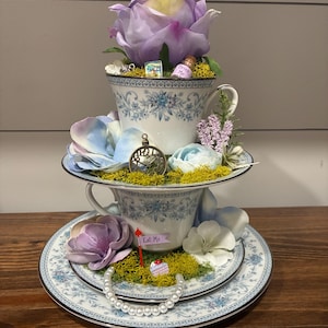 May include: A two-tiered decorative display made of white porcelain teacups and saucers with blue floral patterns. It features artificial purple and blue flowers, green moss, a miniature clock, a bottle labeled Drink Me, and a sign reading Eat Me.