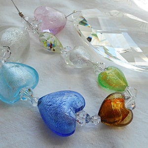 May include: A crystal prism hangs from a string of glass hearts in various colours, including blue, green, gold, and white. The hearts are decorated with silver leaf and have a textured finish.