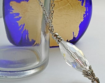 May include: A clear crystal pendant necklace with a silver chain. The pendant is elongated with faceted surfaces, and a silver-toned decorative cap and point. The background includes a blue glass bottle and a gold-colored shape.