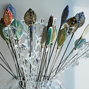 May include: A collection of decorative hat pins in a clear glass vase. The pins feature ornate, leaf-shaped toppers in various colors, including red, blue, teal, and gold. Clear crystal beads and silver stems add to the elegant design.