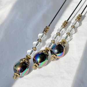 May include: Three decorative hair pins. Each pin has a black stem, clear crystal beads, and a large iridescent bead with gold-coloured accents. The beads have a metallic, oil-slick appearance, reflecting multiple colours.