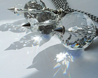 May include: Three crystal teardrop-shaped pendants hang from a silver chain. The pendants are faceted and reflect light, creating rainbows on the surface below.