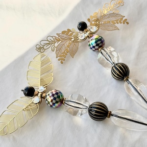 以下が含まれることがあります： 葉と花のアクセントが付いたゴールドトーンのネックレス。ネックレスは、透明、黒、市松模様のビーズが特徴です。デザインには、透明な涙型のビーズと繊細な透かし彫りの葉のデザインが含まれています。