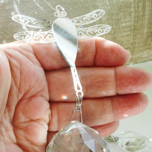May include: A silver-colored dragonfly suncatcher with intricate wing details. The dragonfly is connected to a faceted crystal prism and a smaller crystal pendant. The suncatcher is designed to hang and catch the light.