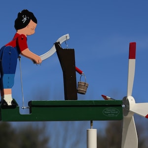 May include: A wooden wind spinner with a girl figure in blue and red clothing operating a well with a bucket. The wind spinner has a white propeller with red and white accents. The base of the wind spinner is green and has the text "Eglien Anleger, Volker Thedens, Since 1993".