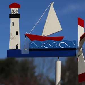 May include: A wooden wind spinner with a red, white, and blue sailboat and lighthouse design. The sailboat has a white sail and is on blue waves. The lighthouse has a red roof and white walls. The wind spinner has a white pole and a red, white, and blue propeller.
