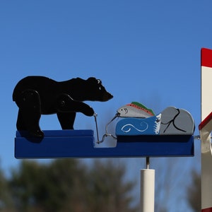 May include: A whimsical wooden weathervane featuring a black bear reaching for a colorful fish. The fish is in a blue boat with white waves. The weathervane has a white and red propeller and a blue base.