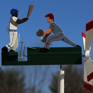 May include: A wooden baseball windsock with two figures, one batting and one catching. The figures are painted in white, blue, red, and gray. The windsock has a green base and a white pole. The windsock has four white blades with red tips.