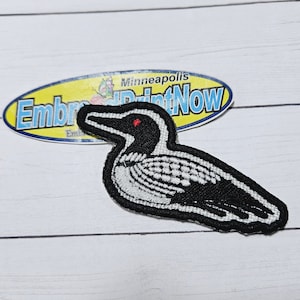 May include: Black and white embroidered patch of a loon bird with a red eye. The patch is on a white wooden surface. The text "Minneapolis PrintNow" is visible on a yellow oval sticker behind the patch.