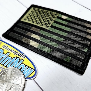 May include: A black and green camouflage American flag patch with a black embroidered star field. The patch is approximately 2.5 inches wide and 3.5 inches long.