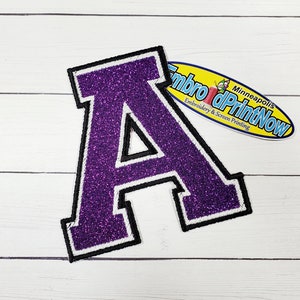 May include: Purple glitter letter A with a white outline and black center. The letter is a felt patch with a stitched edge.