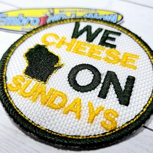 May include: A white, round embroidered patch with a green outline. The patch features the text "WE CHEESE ON SUNDAYS" in yellow and black lettering. The state of Wisconsin is also embroidered in green in the upper left corner.