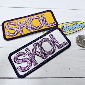 May include: Two embroidered patches with the word "SKOL" in purple and white thread. The top patch is on a yellow background and the bottom patch is on a white background. Both patches have a dark blue border.
