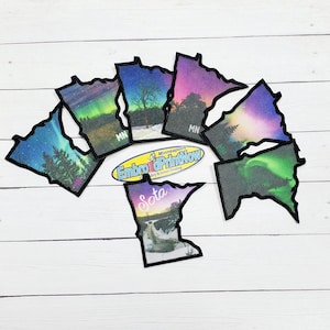 May include: Six embroidered patches shaped like the state of Minnesota. Each patch features a different scenic image of the state, including a waterfall, a forest, and a starry sky. The patches are black with a white outline and the text "MN" and "SOTA" are visible on some of the patches.