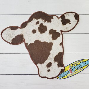 May include: Brown and white cow head patch with a brown stitched outline. The patch is on a white wood background. The text "Embro & PrintNow" is on a blue and yellow oval sticker below the patch.