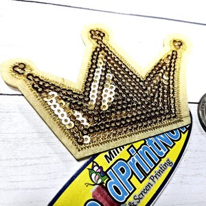 May include: A gold sequined crown patch with a cream-colored border. The crown is next to a US quarter coin. The patch is on top of a sticker with the text "dPrintNow" and a butterfly graphic. The crown is a decorative item.