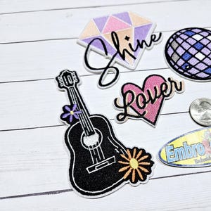 May include: A set of five iron-on patches featuring a pink heart with the word "Lover", a purple and blue disco ball, a purple, pink, and white diamond with the word "Shine", a black acoustic guitar with a purple flower, and a yellow and orange daisy. The patches are on a white wooden surface.