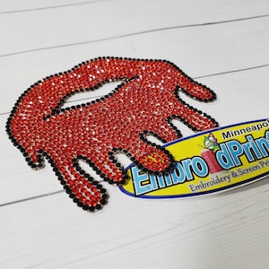 May include: Red rhinestone iron-on patch in the shape of a dripping hand with black outline. The patch is next to a yellow and blue oval sticker with the text "Embro dPrintNow Embroidery & Screen Printing Minneapolis".
