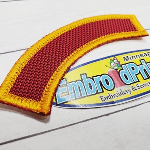 May include: A curved patch in red and yellow with a textured surface. The patch has a yellow border and is made of woven fabric. The text "Embro dPrintNow Embroidery & Screen Printing" is visible on a yellow and white label in the background.