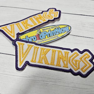 May include: Embroidered patches with the word "VIKINGS" in yellow and white, outlined in purple. The patches are on a light wood grain surface. A sticker with the text "embro dPrintNow" is also visible.