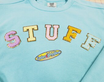 May include: A light blue sweatshirt with the word "STUFF" spelled out in large, colorful, felt letters with gold trim. The letters are sewn on the front of the sweatshirt. A small yellow oval tag with the text "Embro & PrintNow" is sewn on the bottom of the sweatshirt.