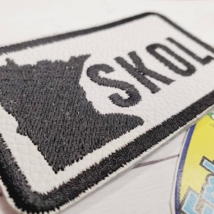 May include: White leather patch with black embroidered outline of the state of Minnesota and the word "SKOL" in black embroidery.