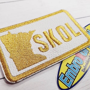 May include: Gold embroidered patch with the word "SKOL" and an outline of the state of Minnesota. The patch is on a white background with a gold border.