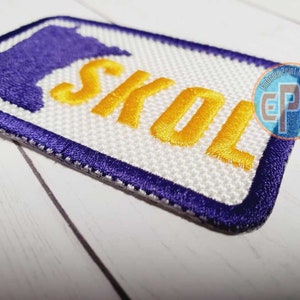 May include: A white embroidered patch with a purple outline and the word "SKOL" in yellow lettering. The patch features a purple outline of the state of Minnesota.