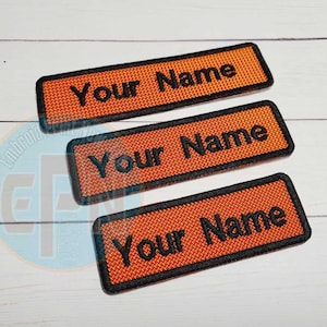 May include: Three rectangular orange patches with black borders and the text "Your Name" embroidered in black. The patches have a textured appearance and are arranged on a white wooden surface.