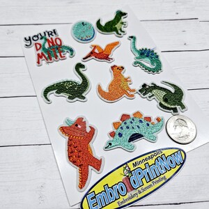 May include: A sheet of iron-on patches featuring various dinosaur designs, including a green dinosaur with the text "You're Dino-mite!" in orange and black. The patches are on a white background with a small business logo in the bottom right corner.