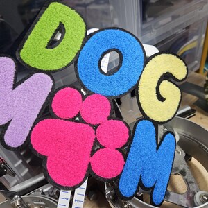 May include: A multi-colored chenille patch featuring the text DOG MOM with a bright pink paw print replacing the letter O. The letters are rendered in lime green, blue, yellow, lavender, and blue with a black border, creating a textured design.