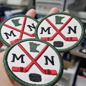 May include: Three embroidered patches with a white background, green border, and the letters "MN". Each patch features crossed red hockey sticks, a green outline of Minnesota, and a black hockey puck. The patches are round.