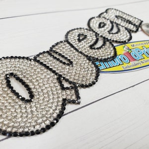 May include: A rhinestone iron-on patch that spells out the word "Queen" in all capital letters. The rhinestones are clear and the letters are outlined in black.