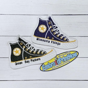 May include: Two embroidered patches of Converse sneakers, one in green and black with the text "Green Bay Packers" and the other in purple and white with the text "Minnesota Vikings".