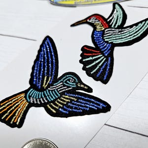 May include: Two embroidered hummingbird patches. One is blue, green, and orange. The other is blue, green, and red.