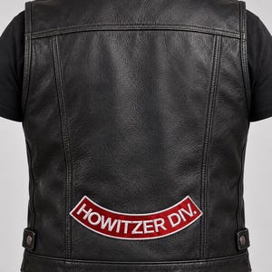 May include: A black leather vest featuring a curved patch on the lower back with a red background and white border. The patch displays the white embroidered text "HOWITZER DIV." The vest has vertical seam detailing and side adjustment tabs.