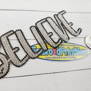 May include: A close-up of a rhinestone iron-on patch that says "BELIEVE" in a stylized font. The patch is made of clear rhinestones with black outlines. The patch is about the size of a US quarter.