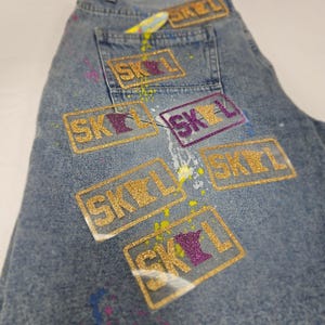 May include: Light blue denim jeans with multiple gold glitter "SKOL" patches. Some patches have a purple glitter design. The jeans have paint splatters in yellow, pink, and blue.
