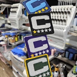 May include: Four embroidered patches in blue, black, purple, and green, each with a white "C" and star accents. The patches are square with a stitched border. The background shows embroidery machines.