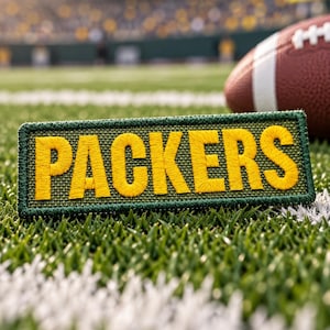 May include: A rectangular embroidered patch featuring the word PACKERS in bold yellow thread on a dark green background with a matching green border, positioned on a green turf field next to a brown leather American football with white laces.