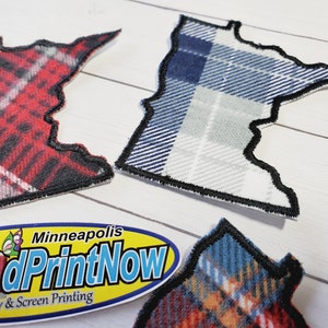 May include: Three Minnesota-shaped patches with different plaid patterns. The patches are made of fabric and have a black stitched border. The patches are on a white wooden surface. The text "Minneapolis PrintNow Embroidery & Screen Printing" is on a yellow oval sticker.