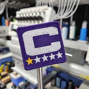 May include: A square purple patch with a white embroidered letter "C" and five stars. The patch has a yellow star on the left side. The patch is held by a metal rod, with an embroidery machine in the background.