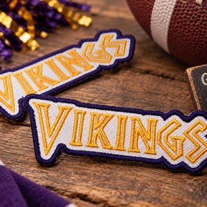 May include: Two embroidered fabric patches featuring the word VIKINGS in yellow block lettering with a white background and a dark purple border. The patches are rectangular with a slight angular shape, showcasing detailed stitching throughout the design.