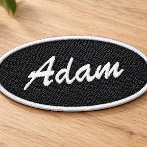 May include: An oval-shaped embroidered patch featuring a black background with a white stitched border. The name Adam is embroidered in the center in a white cursive script font. The patch has a textured fabric surface and clean edges.