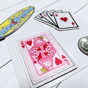 May include: An assortment of embroidered patches and a quarter. The patches include a yellow and blue oval patch with text, playing cards with red hearts, and a pink and red Queen of Hearts card.