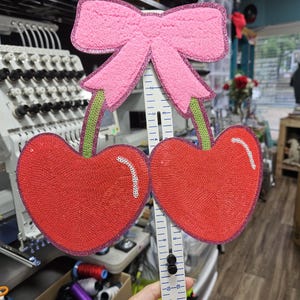 May include: A cherry-shaped patch with red sequins and a pink glittery bow, featuring green stems. A white plastic ruler is positioned vertically behind the patch, showing a height measurement of approximately 8 inches from top to bottom.