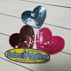 May include: Three heart-shaped sequin patches in blue, red, and pink. The patches are on a white wooden surface. The text "EmbroiderPrintNow" is visible in the bottom of the image.