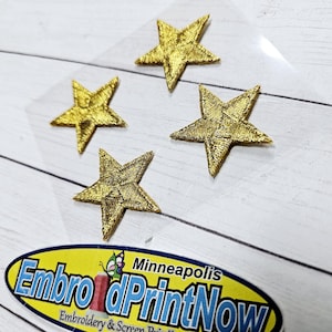 May include: Four gold star-shaped iron-on patches on a white wooden surface. The patches are about 2 inches in diameter.