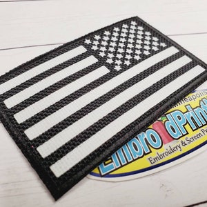May include: Black and white American flag patch with white stitching. The patch is made of a woven fabric and has a black border.