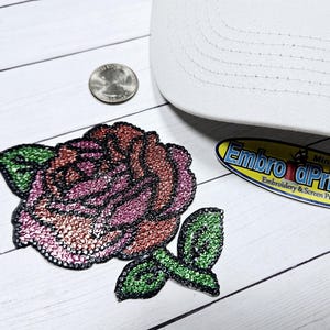 May include: A pink and green rose-shaped iron-on patch with rhinestones. The patch is on a white wooden surface. The text "Embroider & Print Now" is visible in the background.
